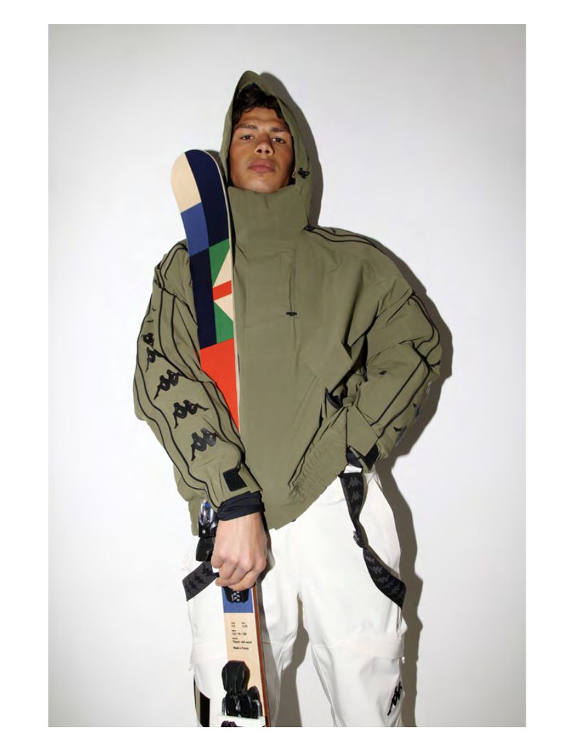 An adult male poses in a hooded jacket and white pants while holding a colorful ski, set against a white backdrop in a studio setting.