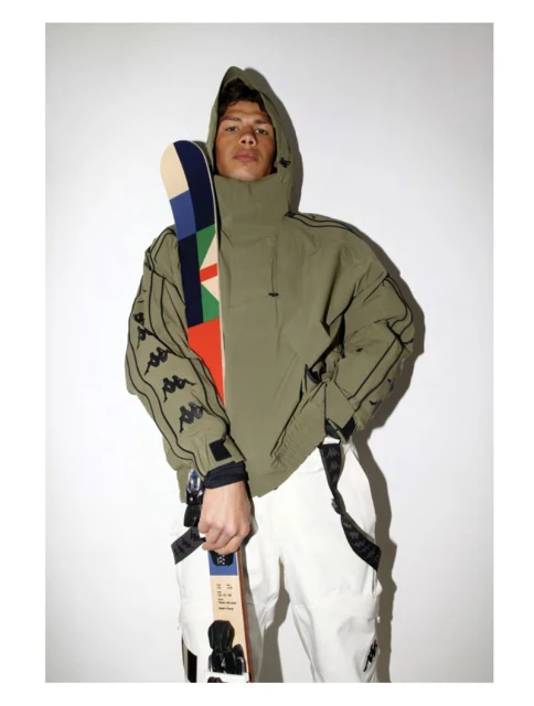 An adult male poses in a hooded jacket and white pants while holding a colorful ski, set against a white backdrop in a studio setting.