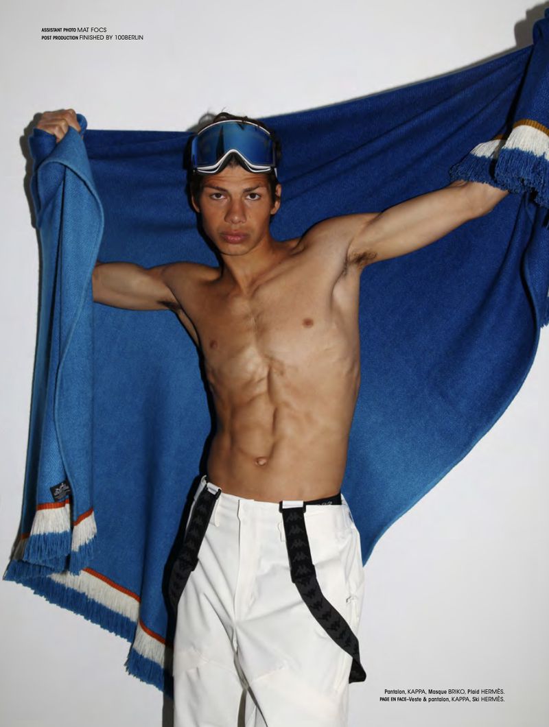 Shirtless man in white pants and ski goggles poses holding a blue and white striped blanket, indoor setting, editorial fashion.