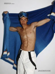 Shirtless male model wearing ski goggles and white trousers holds a large blue fringed blanket against a white background.