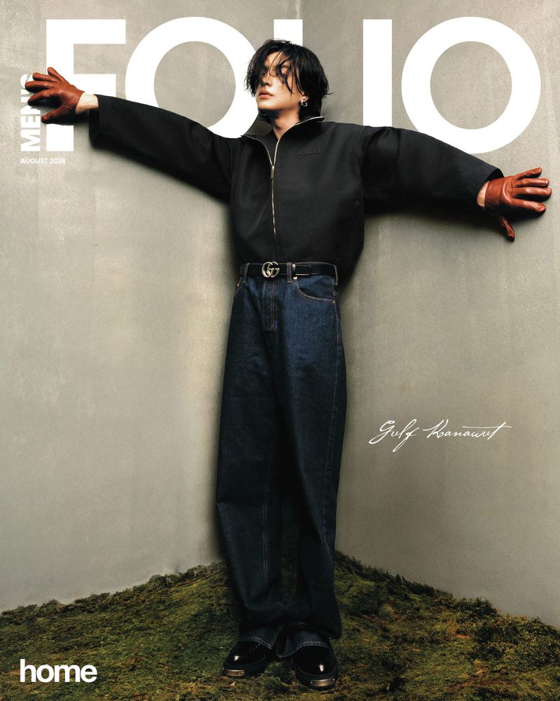 Man in dark jacket and jeans poses on mossy corner for magazine cover, arms outstretched. Editorial fashion shot with muted tones.