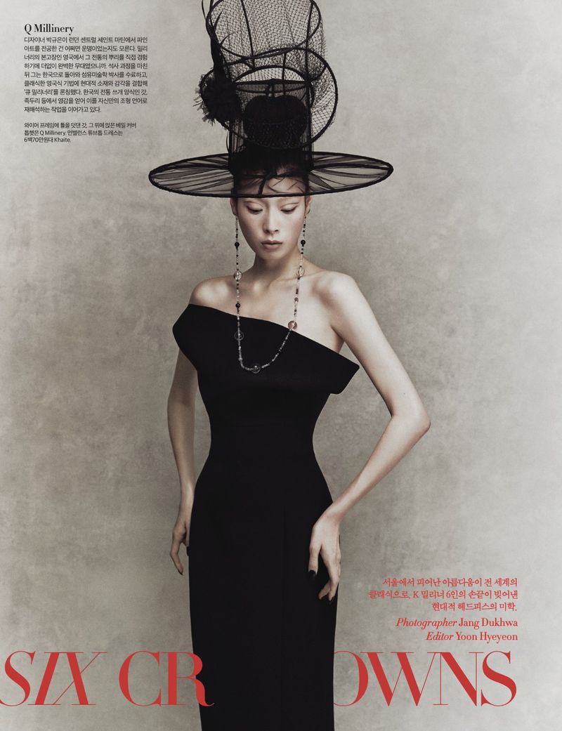 Elegant adult female fashion model poses indoors wearing a black, off-the-shoulder dress, statement necklace, and large decorative hat.