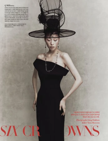 Elegant adult female fashion model poses indoors wearing a black, off-the-shoulder dress, statement necklace, and large decorative hat.