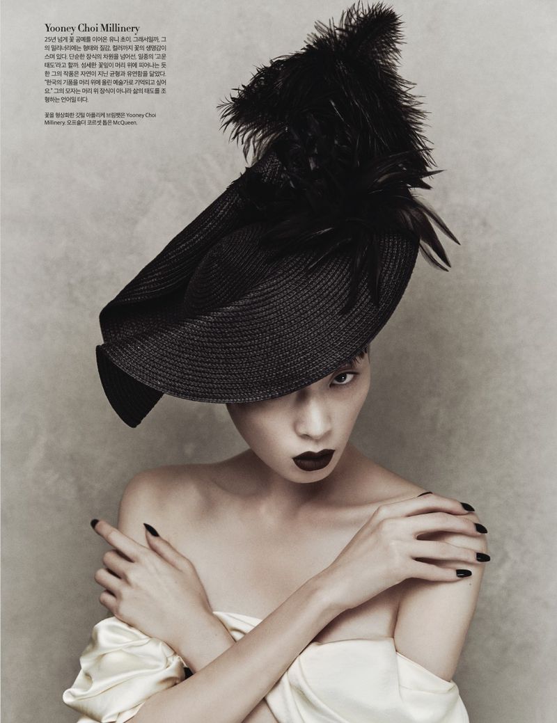 Woman in an editorial fashion shot, wearing a large black hat with feathers, dark lipstick, and pale dress against a textured backdrop.