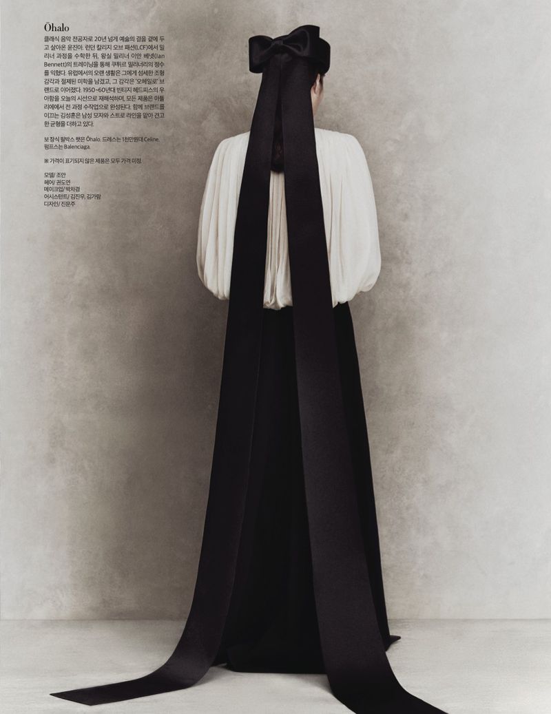 Fashion editorial shot of a woman from the back, wearing a long black gown and a large black ribbon, against a textured grey background.