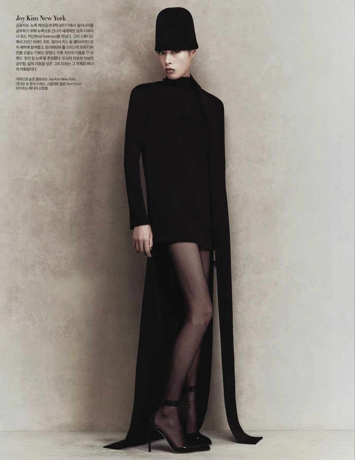 Model in a black dress and tall black hat posing against a textured grey wall for Harper's Bazaar Korea