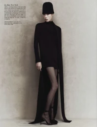 Model in a black dress and tall black hat posing against a textured grey wall for Harper's Bazaar Korea