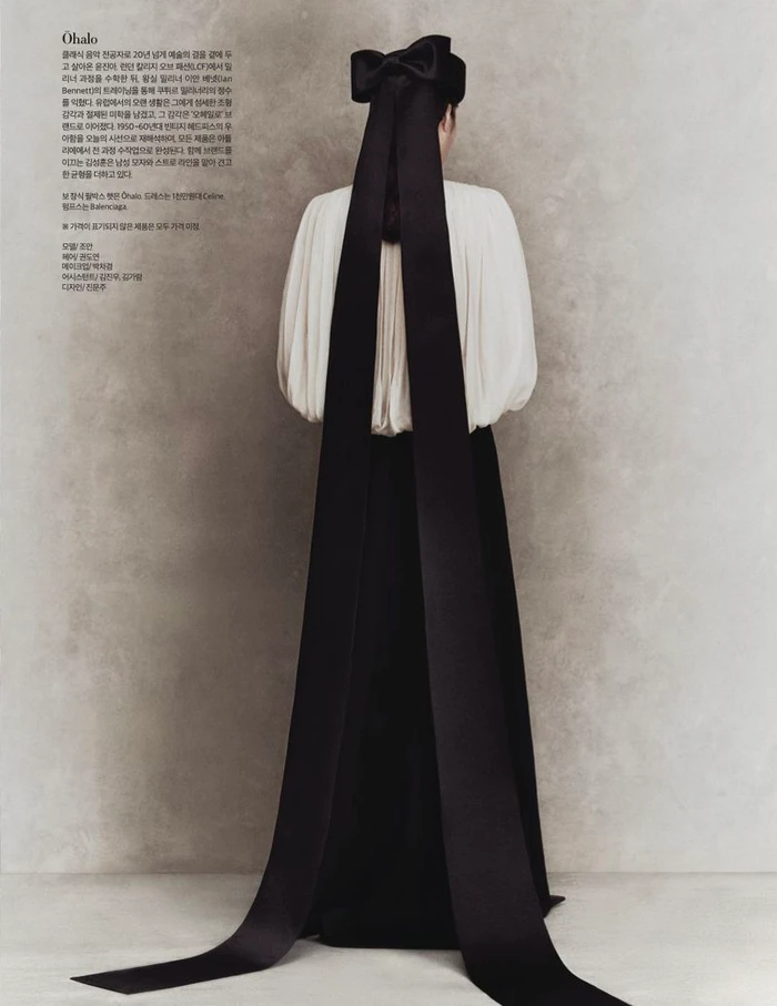 Model seen from behind wearing an oversized black bow with floor-length ribbon tails and a pleated cream blouse against a textured backdrop.