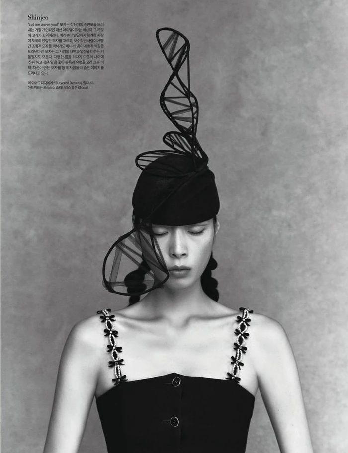 Model Jay Pak poses with eyes closed wearing an elaborate spiral headpiece and black strap top against a textured grey background.