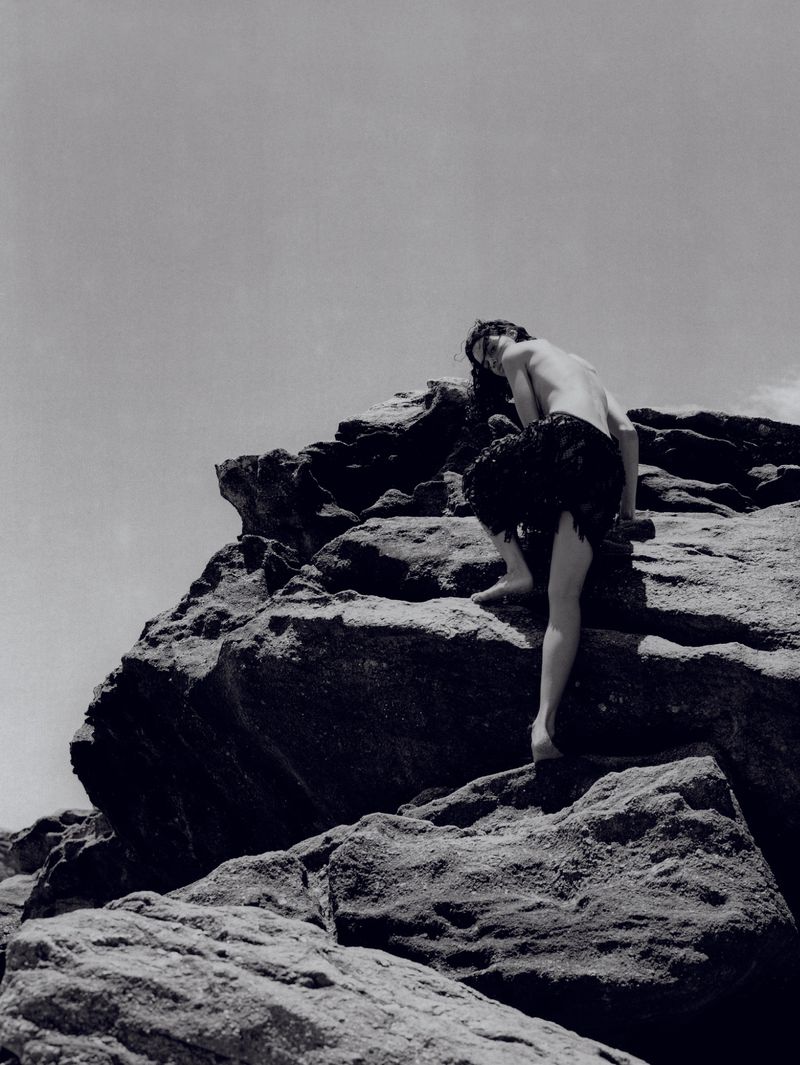 Monochrome shot of a man climbing rocks, wearing a dark textured covering, sky visible in the background, black and white photography.