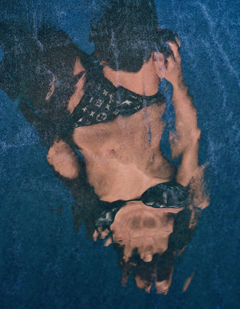 A woman in a bikini stands waist-deep in an indoor pool with blue tile, creating a distorted underwater view with shimmering reflections.