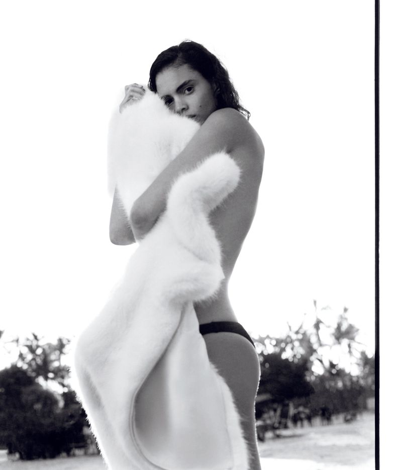 Monochrome portrait of a person hugging a white faux fur coat outdoors, against a bright sky and a palm tree-lined horizon in the background.