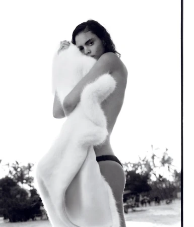 Monochrome portrait of a person hugging a white faux fur coat outdoors, against a bright sky and a palm tree-lined horizon in the background.