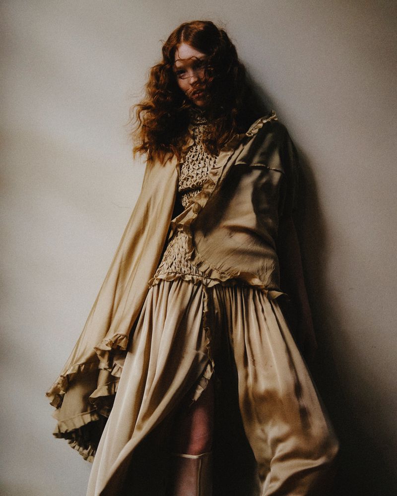 A woman with curly red hair poses in a gold ruffled dress and cape in a fashion editorial photo taken in a photo studio.