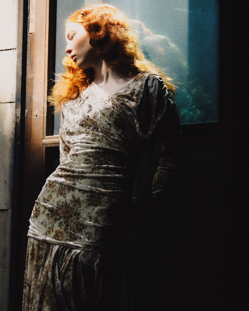 Red-haired woman in a vintage-style dress stands by a window, illuminated by soft natural light creating shadows and highlights on her face and dress.