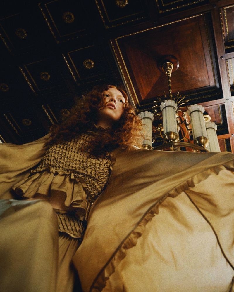 Woman in gold dress stands in ornate room beneath chandelier, captured from a low angle, enhancing the architectural details.