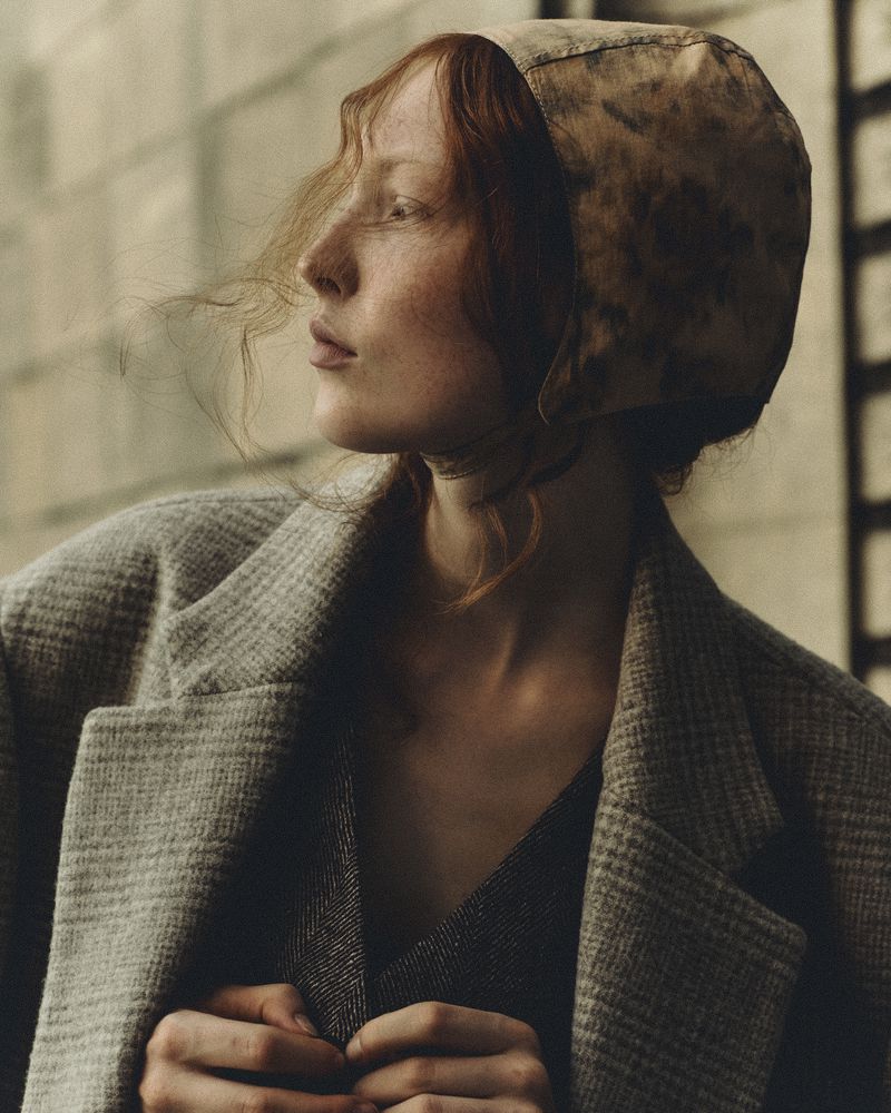 Portrait of a woman with red hair wearing a patterned bonnet and a tweed coat, gazing off into the distance in muted light.