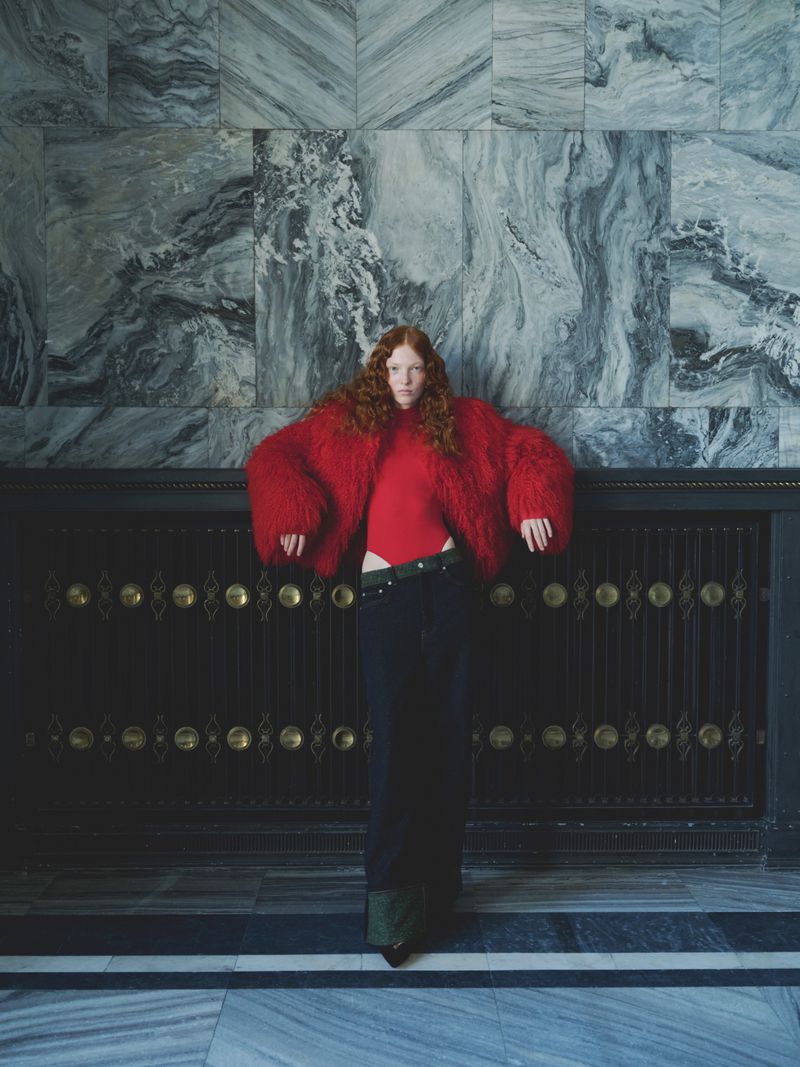 Fashionable redhead woman in a red fur coat and dark skirt poses in front of a decorative marble wall and dark bronze paneling.