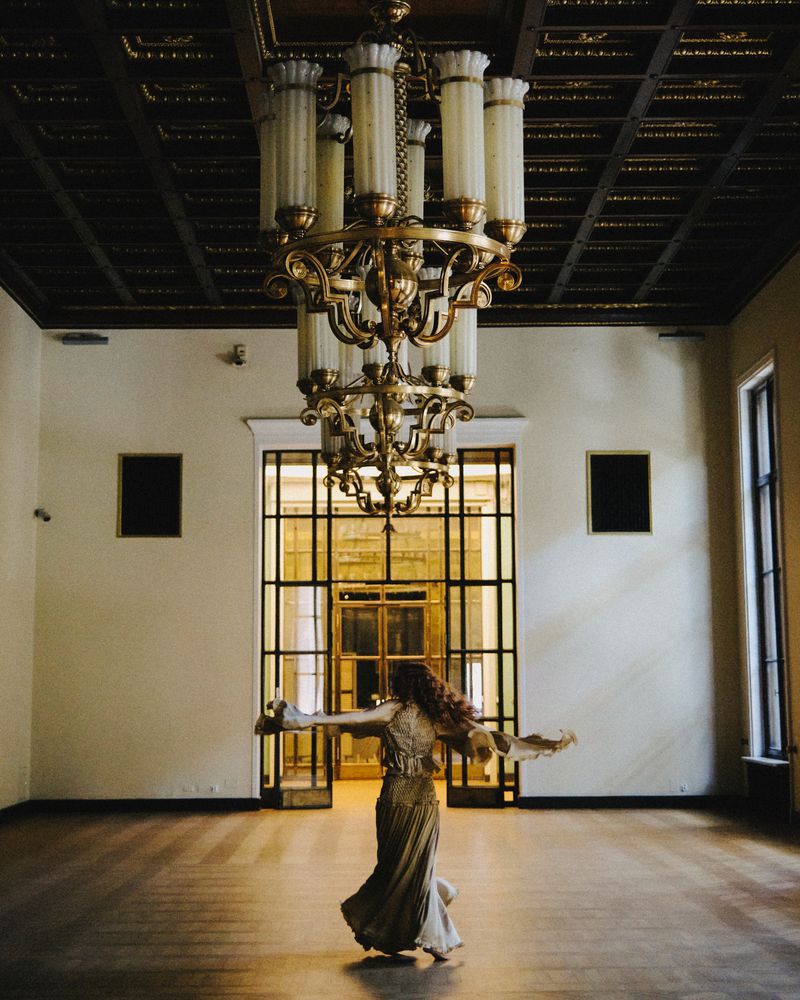 Adult female in flowing gown twirls in ballroom beneath ornate chandelier; large grid door lets in light.