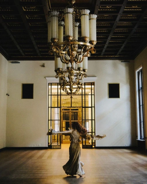 Adult female in flowing gown twirls in ballroom beneath ornate chandelier; large grid door lets in light.