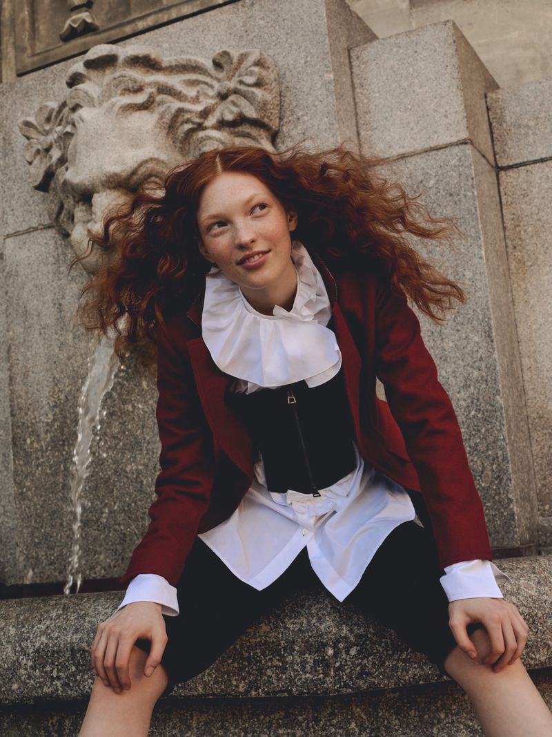 A red-haired woman in a red jacket leans against a stone fountain with a lion head, posing with a relaxed expression and stylish attire.