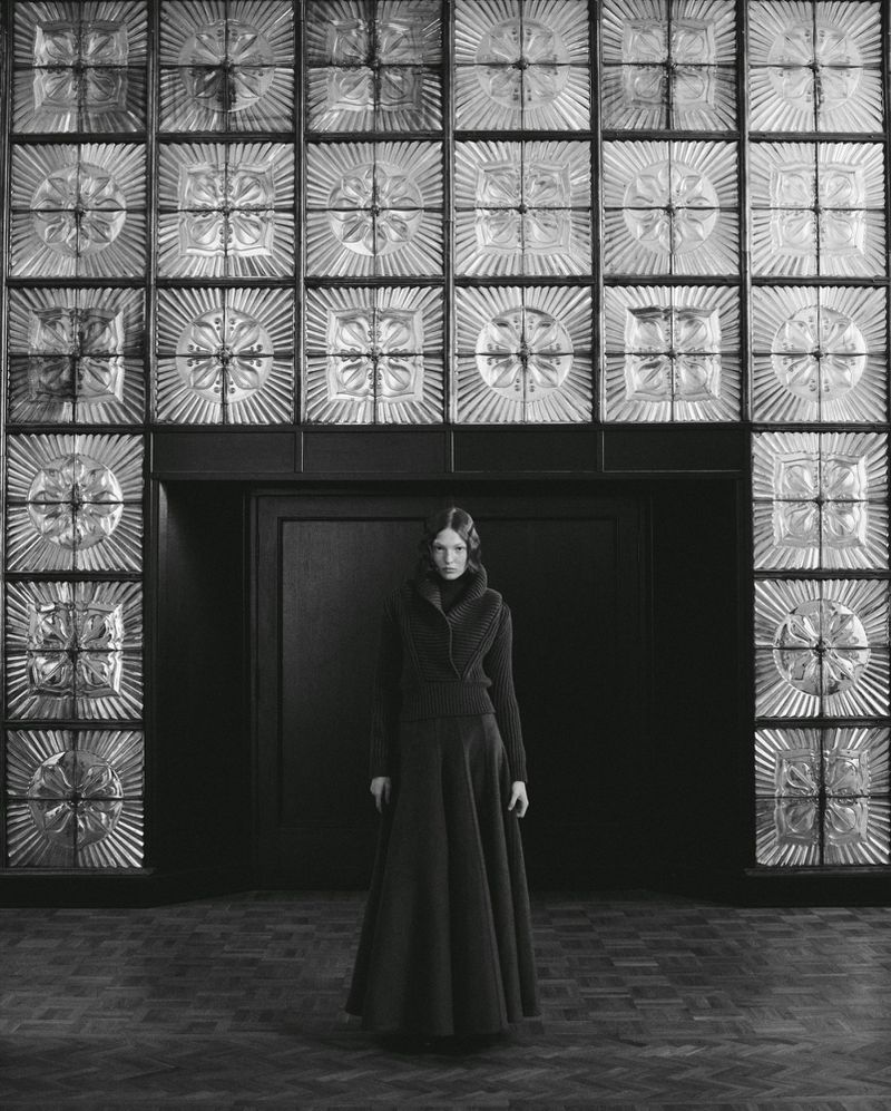 A woman in a long, dark dress stands in a room with decorative glass block walls, captured in a monochromatic, full-length portrait.