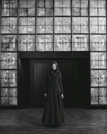 A woman in a long, dark dress stands in a room with decorative glass block walls, captured in a monochromatic, full-length portrait.
