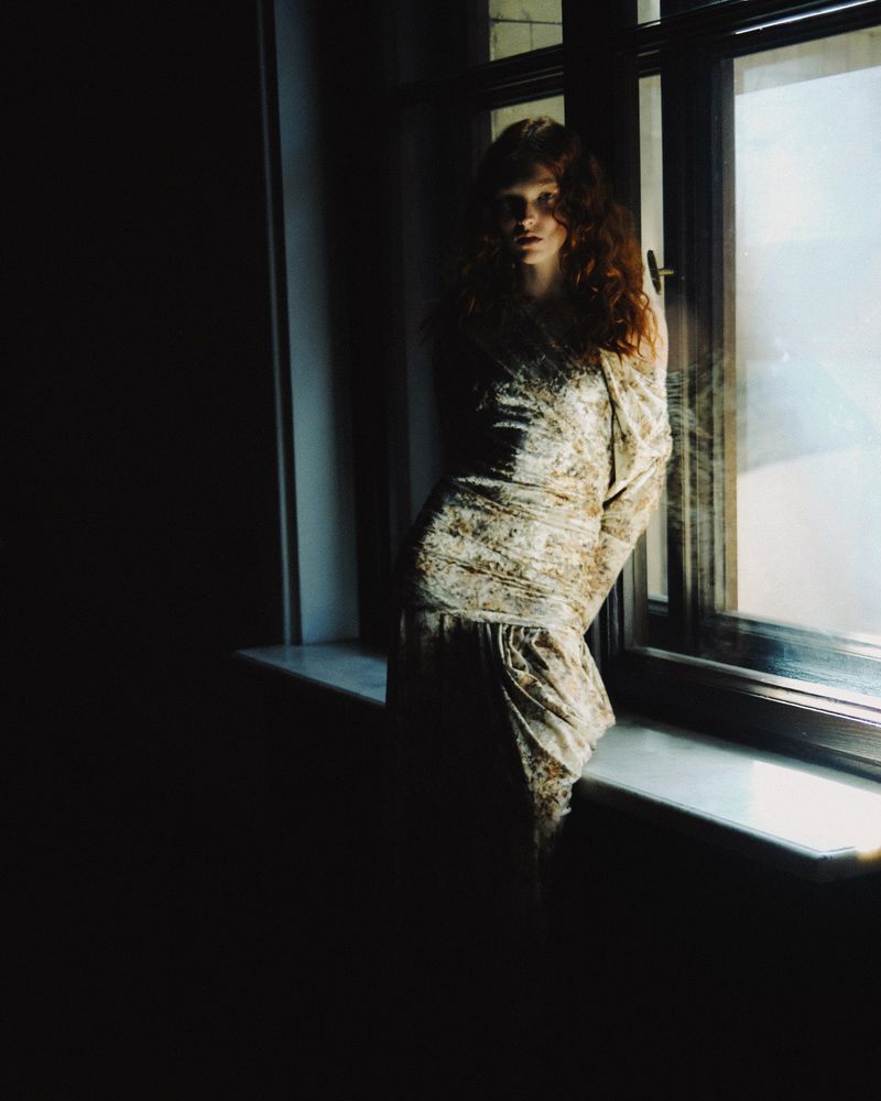 A woman with red curly hair poses near a window in a room with dark lighting, wearing a patterned dress. The window is bright.