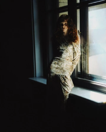 A woman with red curly hair poses near a window in a room with dark lighting, wearing a patterned dress. The window is bright.