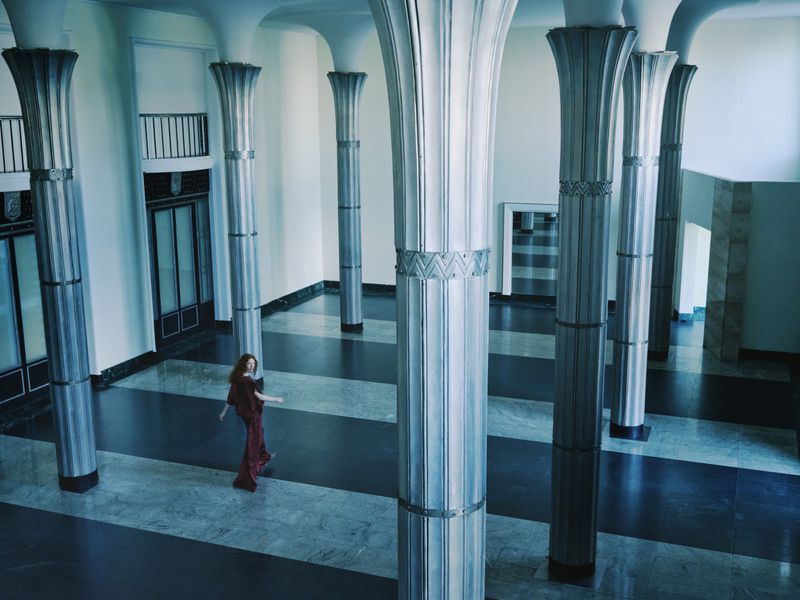 A woman in a red dress walks through an elaborate hall with high ceilings and tall, decorative pillars on a black and white floor.