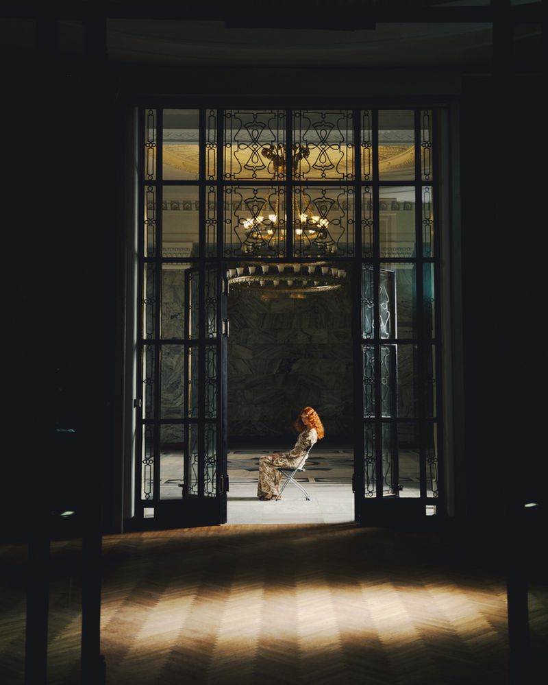 A woman with red hair sits on a chair in a grand, dark room, framed by an open doorway with decorative glass, illuminated by a chandelier.