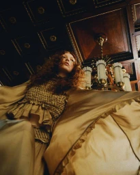 Model with long curly red hair wearing a gold silk outfit, photographed from a dramatic low angle inside an ornate historic hall