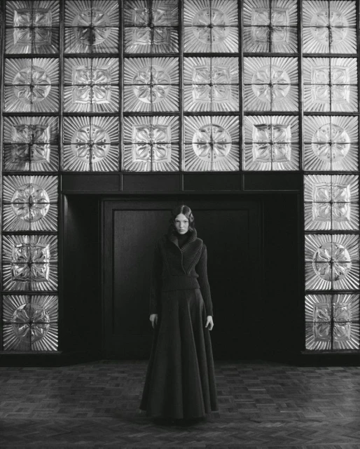 A model wearing a dark long skirt and sweater standing in front of a large patterned glass wall for Elle Poland.