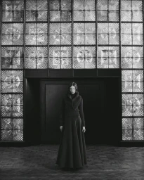 A model wearing a dark long skirt and sweater standing in front of a large patterned glass wall for Elle Poland.