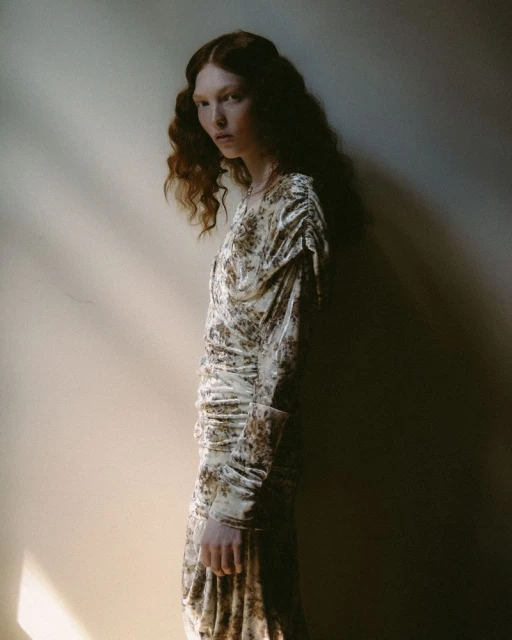 Model with long wavy red hair wearing a patterned dress standing against a plain interior wall in soft directional light