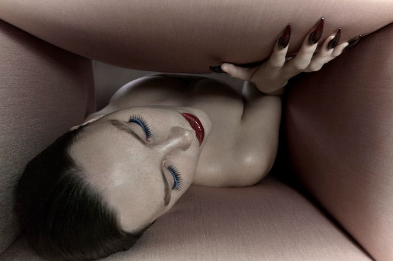 Artistic shot of a woman's face and hand, long nails, striking red lipstick, and blue eyelashes, partially obscured by a pink surface.
