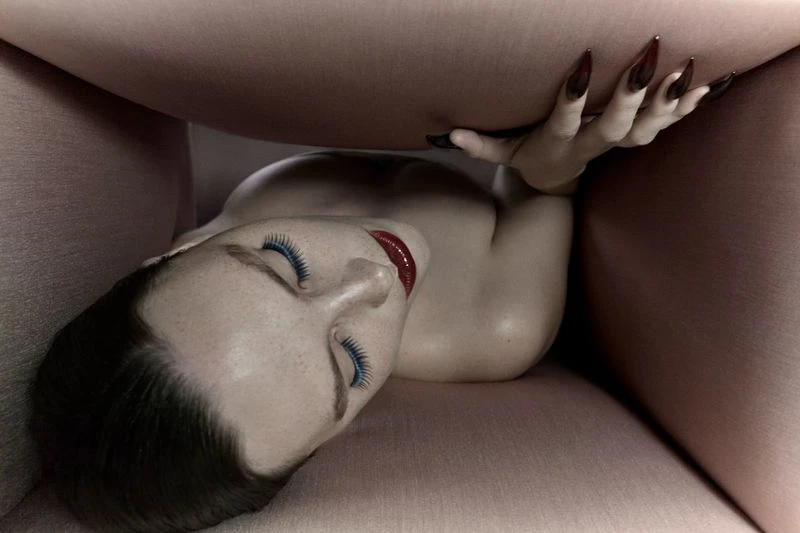 Artistic shot of a woman's face and hand, long nails, striking red lipstick, and blue eyelashes, partially obscured by a pink surface.