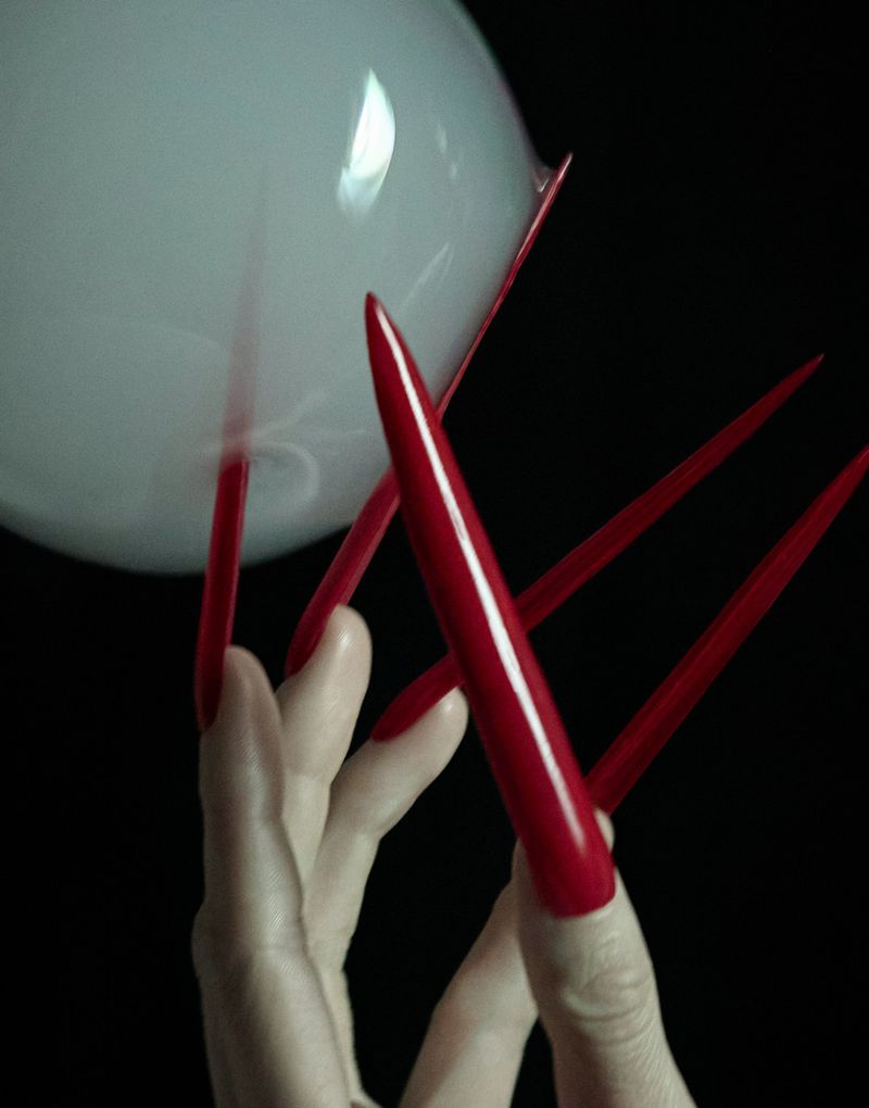Close-up on hand with long, glossy red stiletto nails reaching toward a white, opaque, spherical object against a dark background.