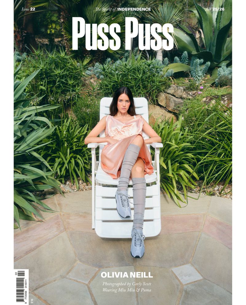 Magazine cover features a woman in a peach dress and knee-high socks sitting on a white chair, surrounded by lush greenery and stone.