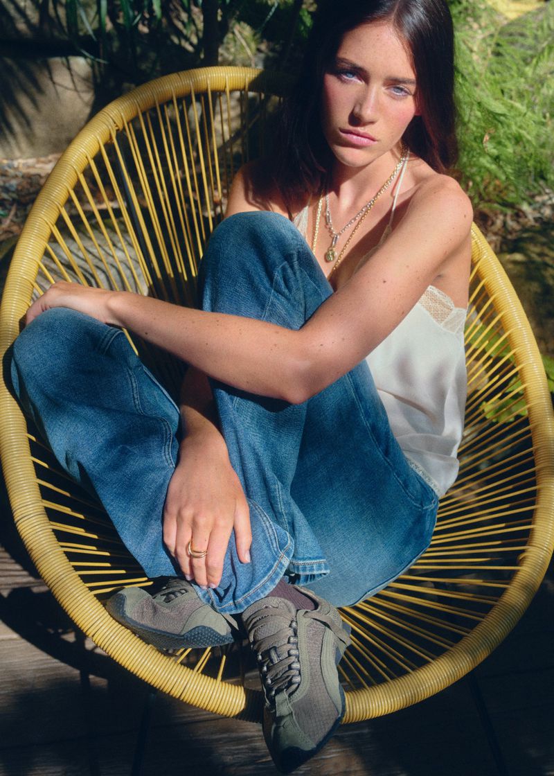 Young woman in casual clothing sits in a yellow wicker chair in a verdant outdoor setting, showcasing a relaxed pose and style.