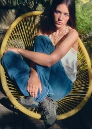 Olivia Neill seated cross-legged in a yellow Acapulco chair wearing blue jeans, a white silk camisole, and sneakers outdoors.