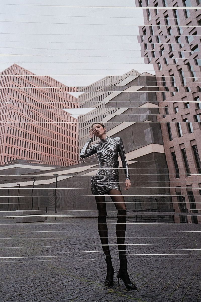 A slender adult woman poses in a metallic dress with an urban architectural background, creating a fashion-editorial image.