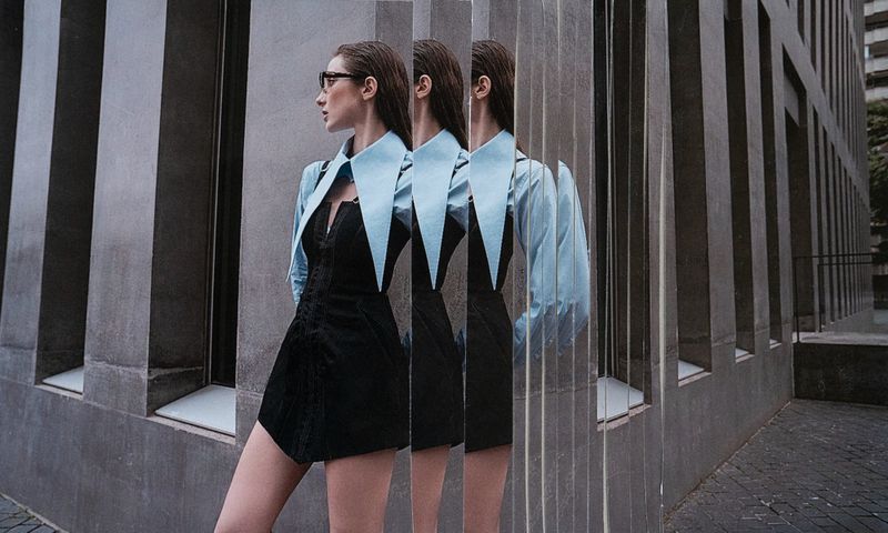 Woman in black dress and light blue collared shirt stands in front of a modern building with repeating, shifted reflections.