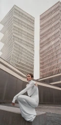 Woman in long white turtleneck dress crouching on a stone wall against tall modern architectural buildings.