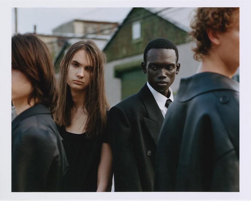 Group of androgynous-looking young adults in formal black clothing. Diverse group poses in front of a weathered building under overcast light.
