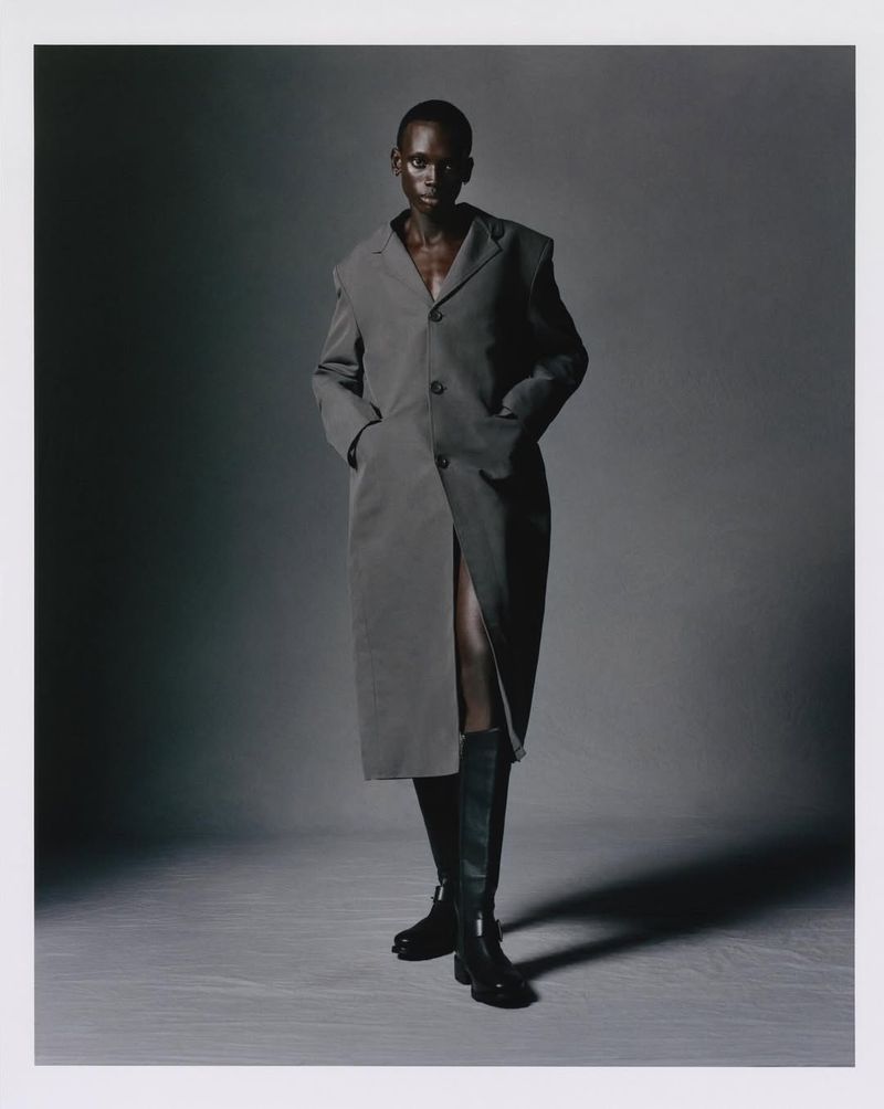 Fashion photo: a statuesque Black woman strikes a pose in a grey coat and knee-high boots against a grey studio backdrop. Style and poise.
