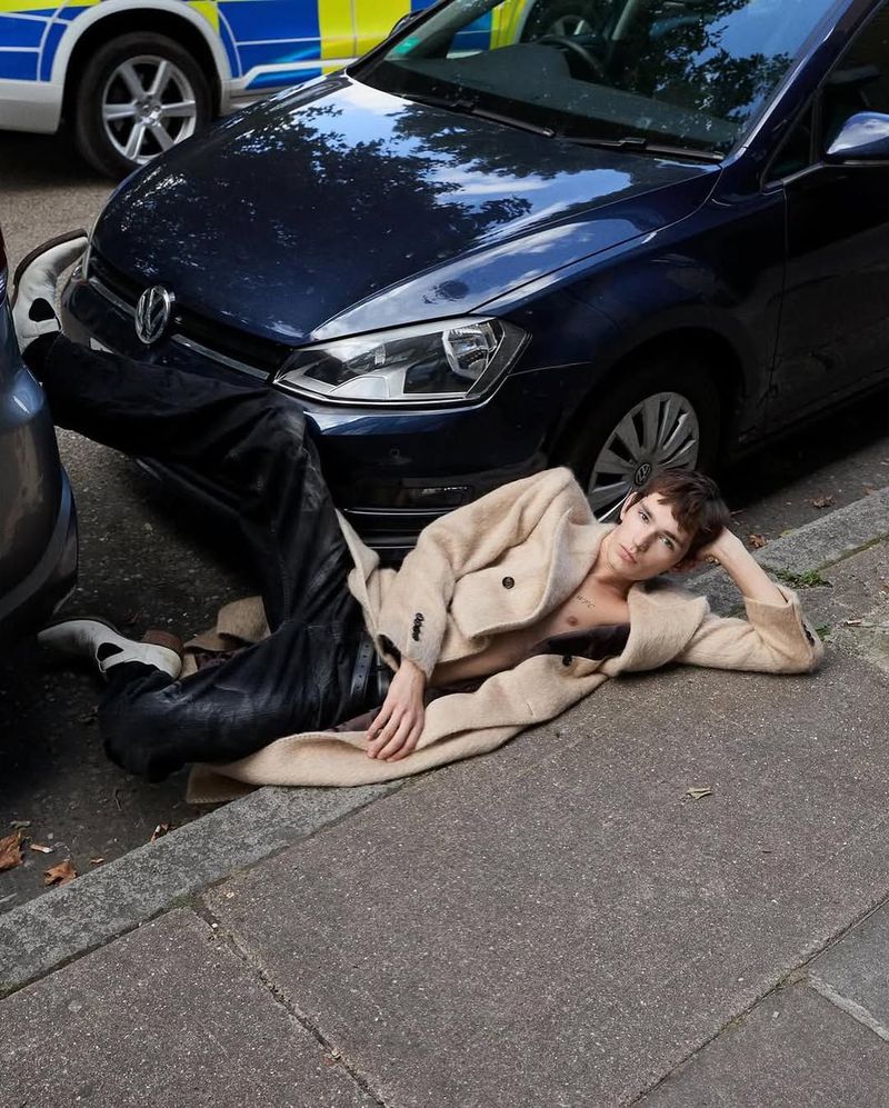 Stylish young adult posing on pavement in unbuttoned coat, near blue car and police vehicle, urban setting, fashion editorial.