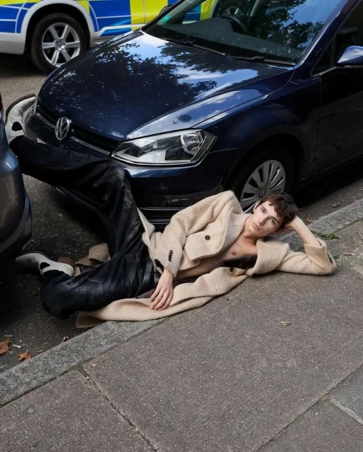 Stylish young adult posing on pavement in unbuttoned coat, near blue car and police vehicle, urban setting, fashion editorial.
