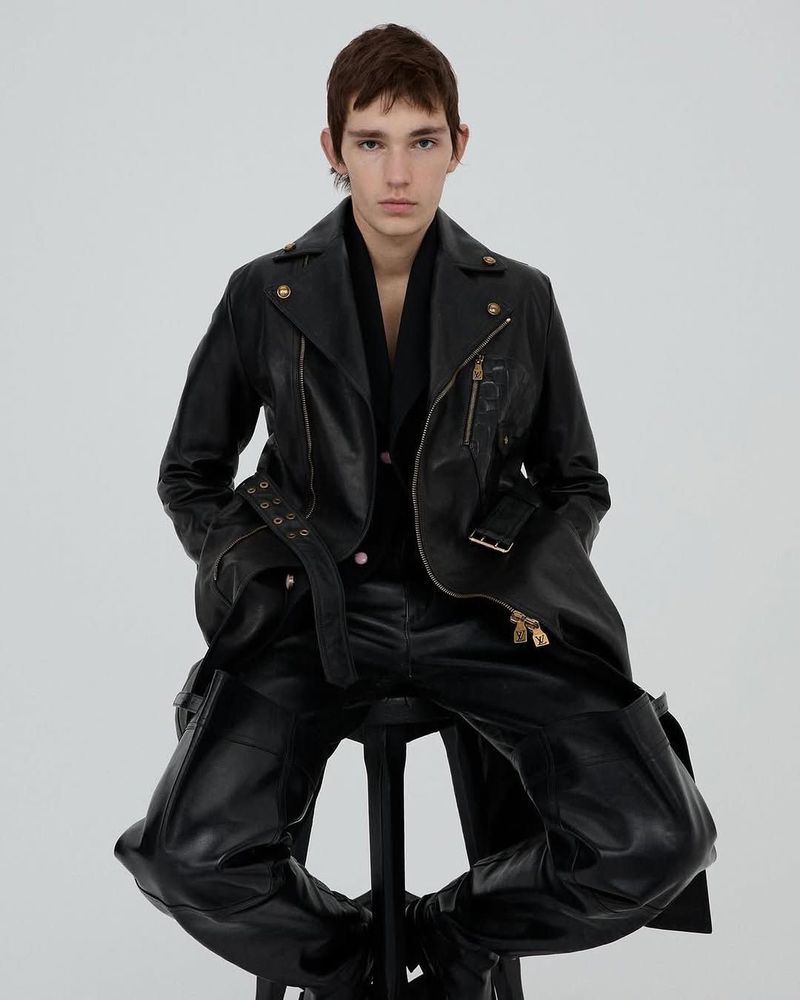 Man in black leather jacket and trousers poses while seated on a stool in a studio with bright, even lighting.