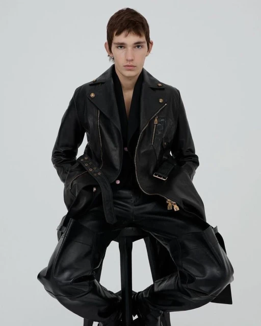 Man in black leather jacket and trousers poses while seated on a stool in a studio with bright, even lighting.